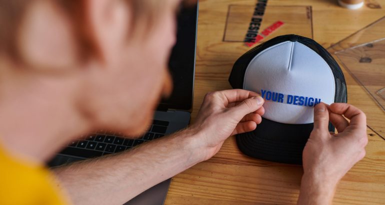 Blurred young designer holding printing layer near snapback while working on project near laptop on table in print studio, hands-on entrepreneurship concept, your design letters
